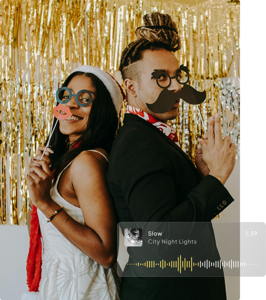 Two people at a fancy dress patty stand back-to-back, posing playfully. They hold up paper props: one with a Santa hat, glasses, and red lips, the other with a mustache, glasses, and tied-up hair. Gold and silver streamers hang in the background.