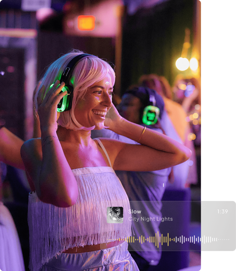A woman with short blonde hair is enjoying a silent disco, wearing glowing headphones. She smiles in a dimly lit room with colourful lights.